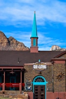 A brick building featuring a tall, bright turquoise spire against a clear blue sky. The front of the building has a sign reading 'Gear & Clothing' for outdoor activities like hiking and climbing. The architecture incorporates elements such as arched windows and exposed brick accents. A mountain range is visible in the background, enhancing the rustic charm of the scene.
