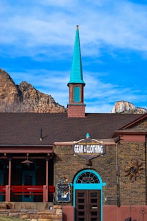A brick building featuring a tall, bright turquoise spire against a clear blue sky. The front of the building has a sign reading 'Gear & Clothing' for outdoor activities like hiking and climbing. The architecture incorporates elements such as arched windows and exposed brick accents. A mountain range is visible in the background, enhancing the rustic charm of the scene.