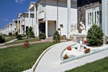 A row of suburban homes with manicured lawns and landscaping, featuring a concrete pathway leading to a white ornamental fountain with red flowers planted nearby. The houses have multiple stories, white or light-colored exteriors, and are arranged uniformly along the street. The sky is clear with a few clouds.
