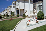 A row of suburban homes with manicured lawns and landscaping, featuring a concrete pathway leading to a white ornamental fountain with red flowers planted nearby. The houses have multiple stories, white or light-colored exteriors, and are arranged uniformly along the street. The sky is clear with a few clouds.