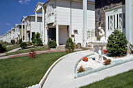 A row of suburban homes with manicured lawns and landscaping, featuring a concrete pathway leading to a white ornamental fountain with red flowers planted nearby. The houses have multiple stories, white or light-colored exteriors, and are arranged uniformly along the street. The sky is clear with a few clouds.