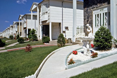 A row of suburban homes with manicured lawns and landscaping, featuring a concrete pathway leading to a white ornamental fountain with red flowers planted nearby. The houses have multiple stories, white or light-colored exteriors, and are arranged uniformly along the street. The sky is clear with a few clouds.