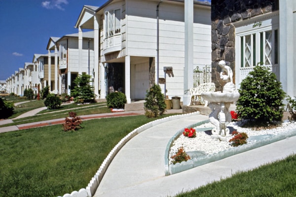 A row of suburban homes with manicured lawns and landscaping, featuring a concrete pathway leading to a white ornamental fountain with red flowers planted nearby. The houses have multiple stories, white or light-colored exteriors, and are arranged uniformly along the street. The sky is clear with a few clouds.