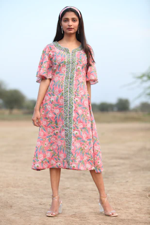 A stylish woman wearing a flowing blush pink dress paired with elegant nude heels, walking confidently in soft natural light.