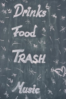 Words such as Drinks, Food, Trash, and Music are written in white chalk on a dark chalkboard. Various doodles and symbols, like stars and a heart, surround the text.