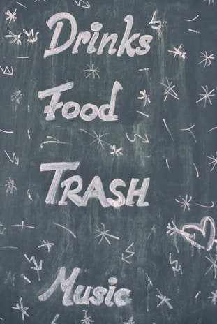Words such as Drinks, Food, Trash, and Music are written in white chalk on a dark chalkboard. Various doodles and symbols, like stars and a heart, surround the text.