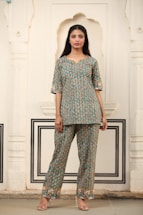 A woman stands in traditional attire featuring a patterned kurta and pants, set against an intricately designed wall with arches and decorative moldings. The outfit has a blue-green base with orange and cream accents. She is wearing transparent high-heeled footwear.