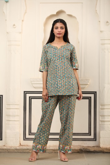 A woman stands in traditional attire featuring a patterned kurta and pants, set against an intricately designed wall with arches and decorative moldings. The outfit has a blue-green base with orange and cream accents. She is wearing transparent high-heeled footwear.
