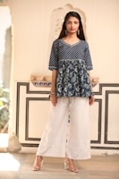 A woman is wearing a blue patterned top with floral designs paired with white wide-legged embroidered pants. She is standing in a stylish, well-lit indoor setting with ornate architectural details in the background. The woman has long, dark hair, and is wearing earrings and clear heels.