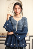 A person with long dark hair is wearing a traditional blue Indian outfit adorned with intricate silver embroidery. The attire includes a top with wide sleeves and floral patterns. Large silver earrings and a matching necklace accessorize the look. The background features an ornate architectural pattern.
