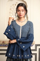 A person with long dark hair is wearing a traditional blue Indian outfit adorned with intricate silver embroidery. The attire includes a top with wide sleeves and floral patterns. Large silver earrings and a matching necklace accessorize the look. The background features an ornate architectural pattern.