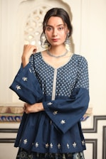 A person with long dark hair is wearing a traditional blue Indian outfit adorned with intricate silver embroidery. The attire includes a top with wide sleeves and floral patterns. Large silver earrings and a matching necklace accessorize the look. The background features an ornate architectural pattern.