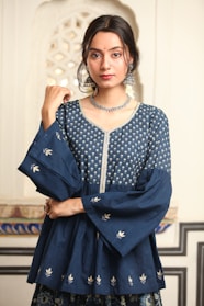A person with long dark hair is wearing a traditional blue Indian outfit adorned with intricate silver embroidery. The attire includes a top with wide sleeves and floral patterns. Large silver earrings and a matching necklace accessorize the look. The background features an ornate architectural pattern.
