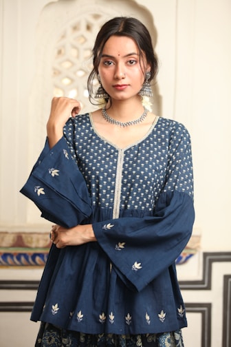 A person with long dark hair is wearing a traditional blue Indian outfit adorned with intricate silver embroidery. The attire includes a top with wide sleeves and floral patterns. Large silver earrings and a matching necklace accessorize the look. The background features an ornate architectural pattern.