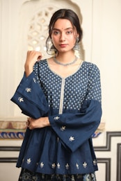 A person with long dark hair is wearing a traditional blue Indian outfit adorned with intricate silver embroidery. The attire includes a top with wide sleeves and floral patterns. Large silver earrings and a matching necklace accessorize the look. The background features an ornate architectural pattern.