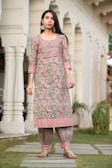 A woman stands in a garden setting, wearing a traditional South Asian outfit with intricate floral patterns. The outfit consists of a long, patterned tunic and matching pants, complemented by large decorative earrings. The background features arched architectural elements and greenery.