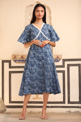 A person stands confidently indoors, wearing a stylish blue floral dress with white lace trim. They have long, dark hair and are accessorized with a choker necklace and a large ring. The background features ornate, traditional architectural details with light-colored walls and intricate designs.