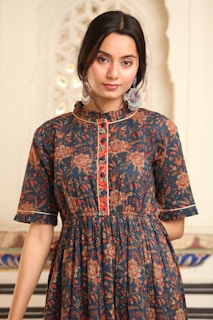 A person is wearing a dark blue dress with intricate floral patterns in shades of red, orange, and brown. The dress has buttons down the front and a ruffled collar. The person has dark hair styled back and is wearing large, ornate earrings with colorful designs. The background is a soft, light color with a decorative pattern visible.