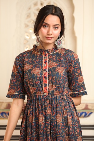 A person is wearing a dark blue dress with intricate floral patterns in shades of red, orange, and brown. The dress has buttons down the front and a ruffled collar. The person has dark hair styled back and is wearing large, ornate earrings with colorful designs. The background is a soft, light color with a decorative pattern visible.
