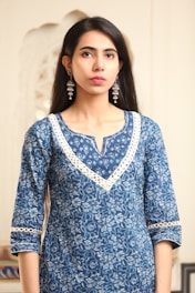 A person is wearing a blue, floral-patterned kurta with detailed white lace trim around the neckline and sleeves. The individual has long, dark hair and is accessorized with long, ornate silver earrings. The background appears to be indoors with soft, neutral tones.