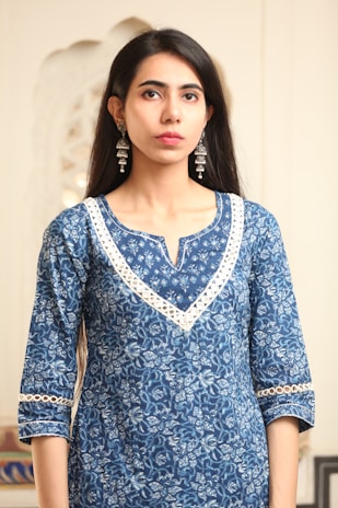 A person is wearing a blue, floral-patterned kurta with detailed white lace trim around the neckline and sleeves. The individual has long, dark hair and is accessorized with long, ornate silver earrings. The background appears to be indoors with soft, neutral tones.
