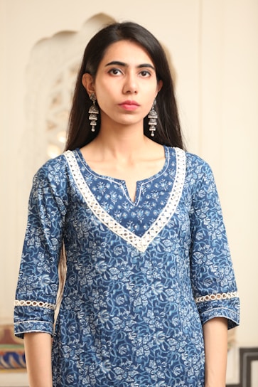 A person is wearing a blue, floral-patterned kurta with detailed white lace trim around the neckline and sleeves. The individual has long, dark hair and is accessorized with long, ornate silver earrings. The background appears to be indoors with soft, neutral tones.