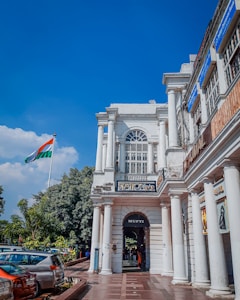 A colonial-style building with tall white columns and arched windows is situated on a bustling street. The Indian national flag flutters prominently in the background against a clear blue sky. Cars are parked along the road, and lush greenery lines the walkway. Several signs and shop banners are visible on the building.