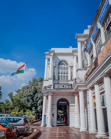 A colonial-style building with tall white columns and arched windows is situated on a bustling street. The Indian national flag flutters prominently in the background against a clear blue sky. Cars are parked along the road, and lush greenery lines the walkway. Several signs and shop banners are visible on the building.