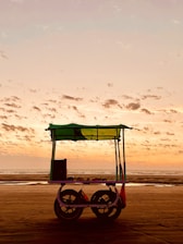 A small cart with a canopy sits on a beach during sunset. The sky is filled with scattered clouds and is painted in soft hues of orange and pink. The cart has two large wheels and seems to be used for selling items, possibly food or drinks.