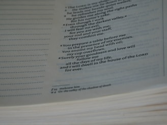 A page from a book with printed text, displaying religious verses. The text is organized in numbered lines, with additional explanatory notes at the bottom of the page. The page appears slightly angled, and the lighting is soft, creating a serene atmosphere.