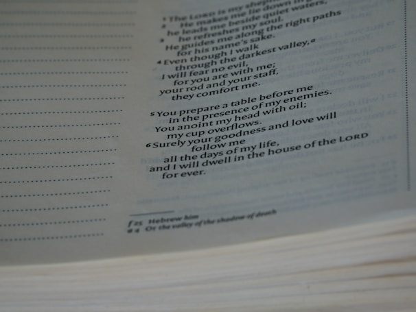 A page from a book with printed text, displaying religious verses. The text is organized in numbered lines, with additional explanatory notes at the bottom of the page. The page appears slightly angled, and the lighting is soft, creating a serene atmosphere.