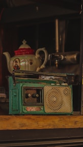 A vintage teal radio with dials and a speaker grill sits prominently on a wooden surface. In the background, there is an ornate teapot with a red lid and decorative images on its sides.