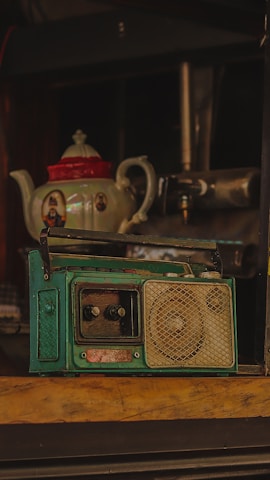 A vintage teal radio with dials and a speaker grill sits prominently on a wooden surface. In the background, there is an ornate teapot with a red lid and decorative images on its sides.