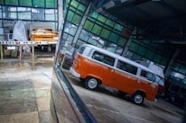 Inside a spacious industrial garage, there is a vintage orange and white van with a distinctive design. The reflections on the shiny floor enhance the vividness of the van's color. In the background, another vehicle is covered with a protective sheet, and various tools and construction materials are scattered throughout the space, indicating ongoing automotive work.