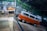 Inside a spacious industrial garage, there is a vintage orange and white van with a distinctive design. The reflections on the shiny floor enhance the vividness of the van's color. In the background, another vehicle is covered with a protective sheet, and various tools and construction materials are scattered throughout the space, indicating ongoing automotive work.