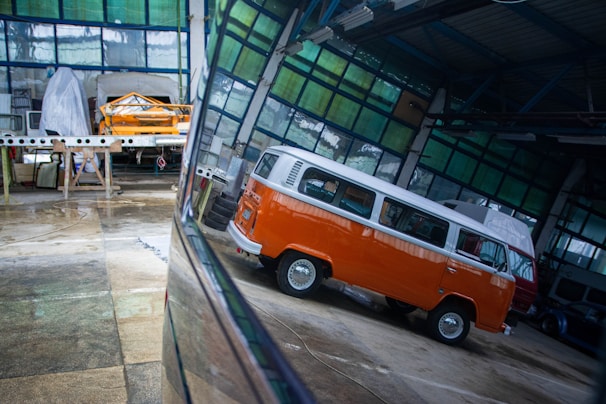 Inside a spacious industrial garage, there is a vintage orange and white van with a distinctive design. The reflections on the shiny floor enhance the vividness of the van's color. In the background, another vehicle is covered with a protective sheet, and various tools and construction materials are scattered throughout the space, indicating ongoing automotive work.