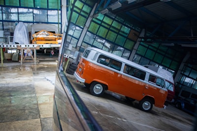 Inside a spacious industrial garage, there is a vintage orange and white van with a distinctive design. The reflections on the shiny floor enhance the vividness of the van's color. In the background, another vehicle is covered with a protective sheet, and various tools and construction materials are scattered throughout the space, indicating ongoing automotive work.
