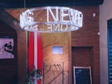 A cozy interior with a large circular neon sign suspended from the ceiling, casting a white glow. A wooden wall with diagonal panels features a mounted surfboard and a small framed picture. To the left, there is a poster of a person dining, and below it is a green plant. A chalkboard with some scribbles is seen on the right.