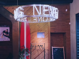 A cozy interior with a large circular neon sign suspended from the ceiling, casting a white glow. A wooden wall with diagonal panels features a mounted surfboard and a small framed picture. To the left, there is a poster of a person dining, and below it is a green plant. A chalkboard with some scribbles is seen on the right.