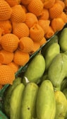 A close-up of a fruit display at a market, showing stacked oranges in orange protective netting on the left side and a pile of green mangoes on the right side.
