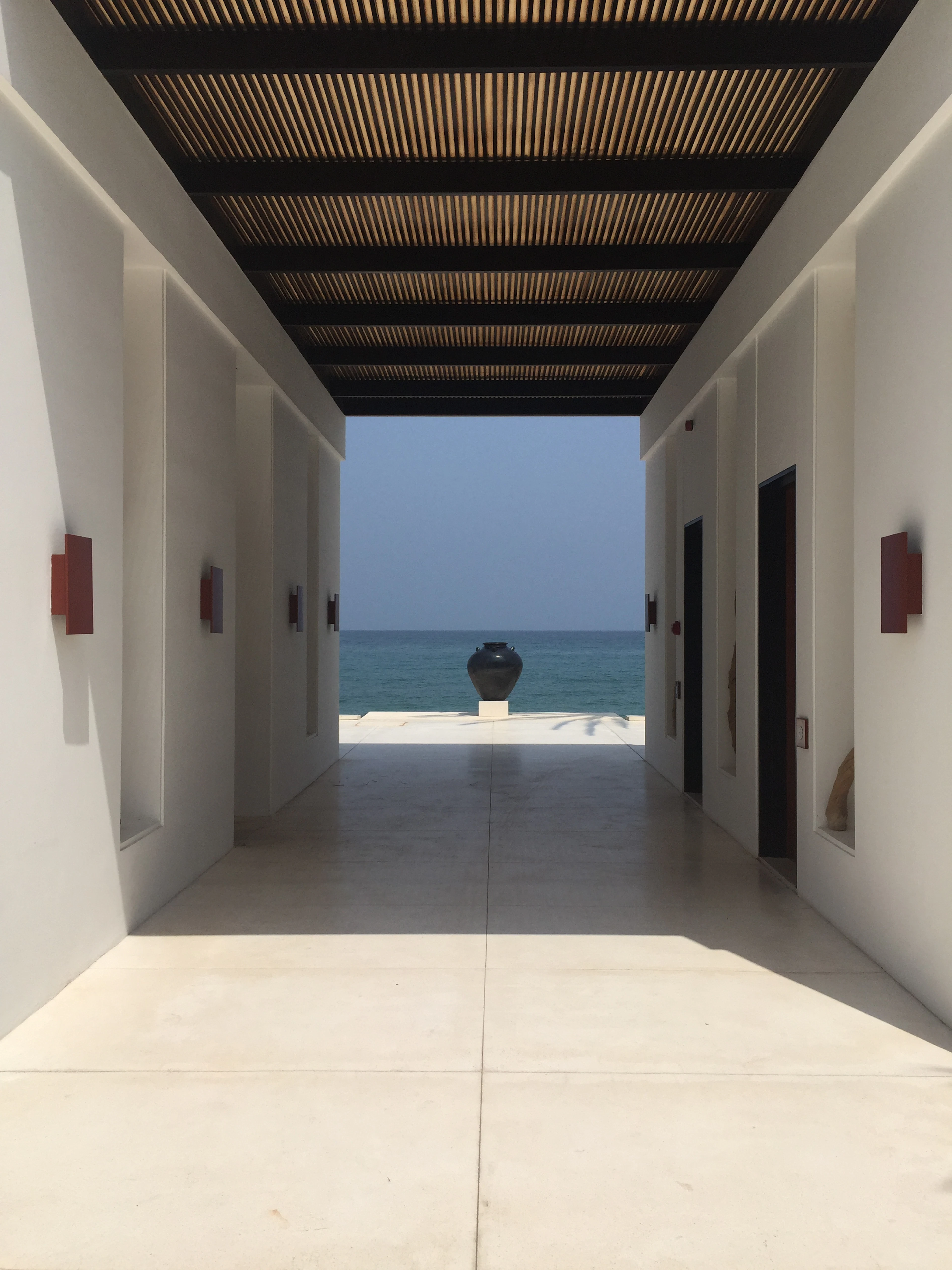 A shaded slatted colonnade at The Chedi Muscat opens onto the turquoise Gulf of Oman, a single dark urn standing at the end of the pale stone walkway.