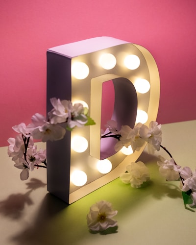 A large, illuminated letter D decorated with white flowers is set against a pastel pink background. The letter has multiple small light bulbs embedded in it, creating a bright and inviting glow. The surrounding flowers give a soft, natural contrast to the structured shape of the letter.