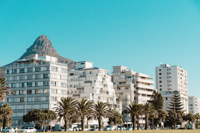 Several modern apartment buildings are lined up under a clear blue sky with a mountain in the background. Palm trees are in front of the buildings, and there are a few cars parked along the street.