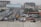A busy urban street with several vehicles including yellow taxis, a white van, and a truck. There are multi-story buildings under construction, and a large billboard over the road with the text 'Stand Out From The Crowd, Advertise Here'. Pedestrians are walking along the sidewalks, and the area is surrounded by various commercial and residential buildings.