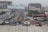 A busy urban street with several vehicles including yellow taxis, a white van, and a truck. There are multi-story buildings under construction, and a large billboard over the road with the text 'Stand Out From The Crowd, Advertise Here'. Pedestrians are walking along the sidewalks, and the area is surrounded by various commercial and residential buildings.