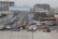 A busy urban street with several vehicles including yellow taxis, a white van, and a truck. There are multi-story buildings under construction, and a large billboard over the road with the text 'Stand Out From The Crowd, Advertise Here'. Pedestrians are walking along the sidewalks, and the area is surrounded by various commercial and residential buildings.