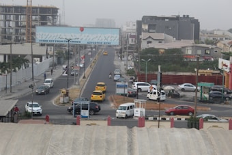 A busy urban street with several vehicles including yellow taxis, a white van, and a truck. There are multi-story buildings under construction, and a large billboard over the road with the text 'Stand Out From The Crowd, Advertise Here'. Pedestrians are walking along the sidewalks, and the area is surrounded by various commercial and residential buildings.