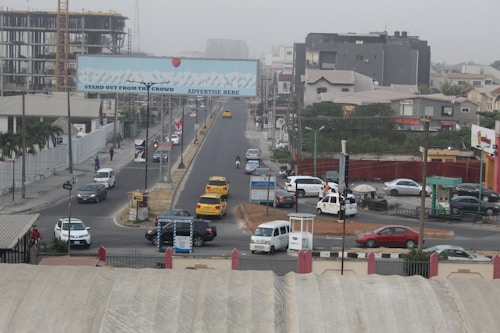 A busy urban street with several vehicles including yellow taxis, a white van, and a truck. There are multi-story buildings under construction, and a large billboard over the road with the text 'Stand Out From The Crowd, Advertise Here'. Pedestrians are walking along the sidewalks, and the area is surrounded by various commercial and residential buildings.
