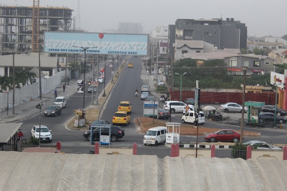 A busy urban street with several vehicles including yellow taxis, a white van, and a truck. There are multi-story buildings under construction, and a large billboard over the road with the text 'Stand Out From The Crowd, Advertise Here'. Pedestrians are walking along the sidewalks, and the area is surrounded by various commercial and residential buildings.