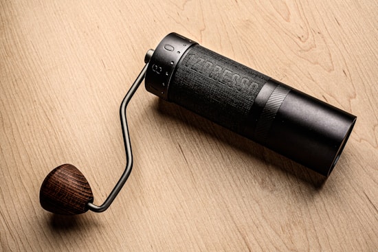 A manual coffee grinder lies on a wooden surface. The grinder features a cylindrical black body with the brand name etched into it and a dark metallic handle leading to a wooden knob. The craftsmanship of the grinder highlights a blend of modern and traditional design elements.